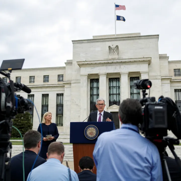 Press briefing in front of the Federal Reserve headquarters in Washington – a speaker stands at a lectern bearing the Fed seal, with journalists' cameras in the foreground.