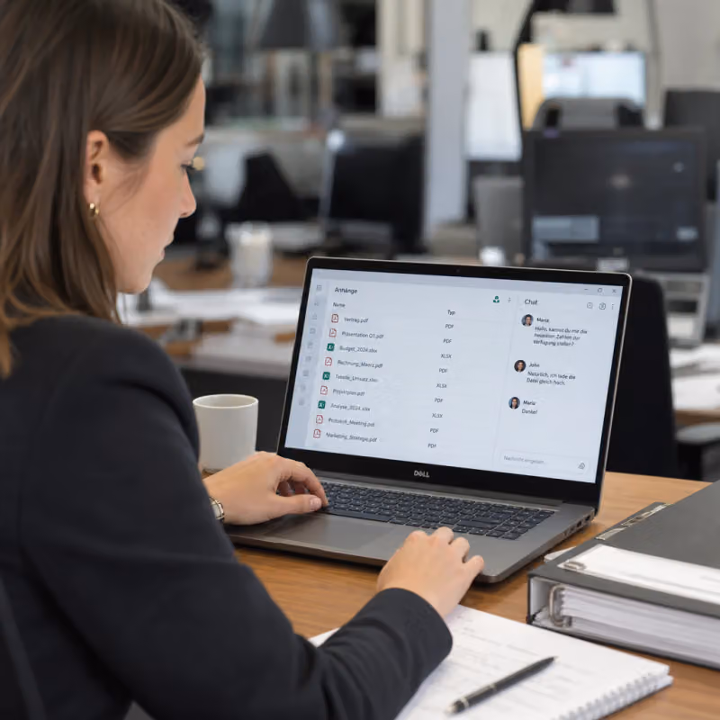 An office worker checks a list of email attachments created by Copilot next to a file folder on the desk using a laptop.