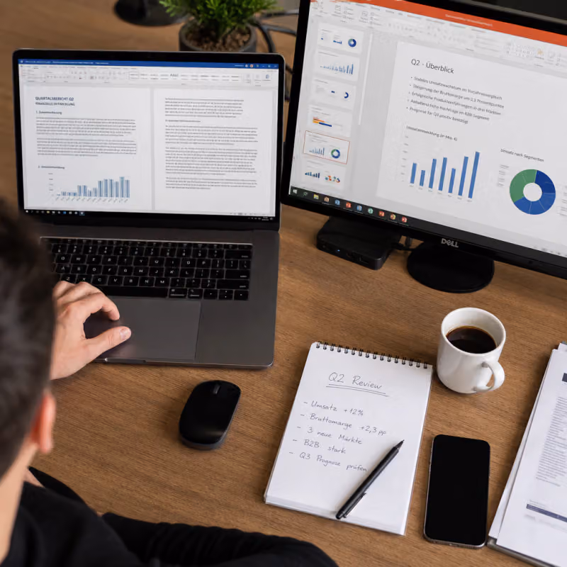Desk with laptop, on whose screen a Word document with a quarterly report is visible, next to a second monitor with an overview of PowerPoint slides.