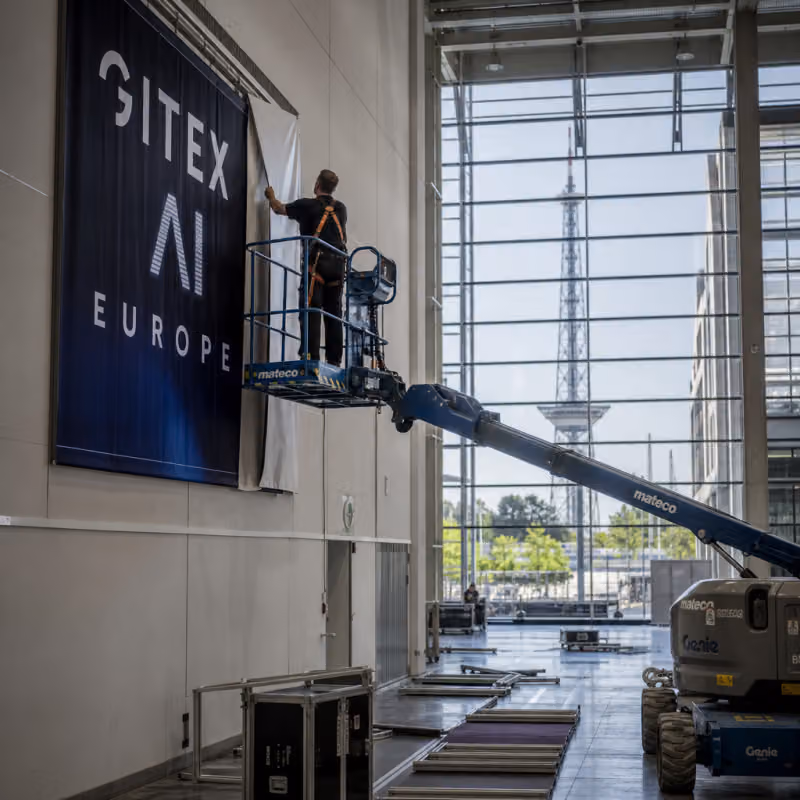 Photorealistic depiction: in an empty Berlin exhibition hall, a technician on a lift removes a banner reading "GITEX AI Europe"; the Berlin radio tower is visible in the background.