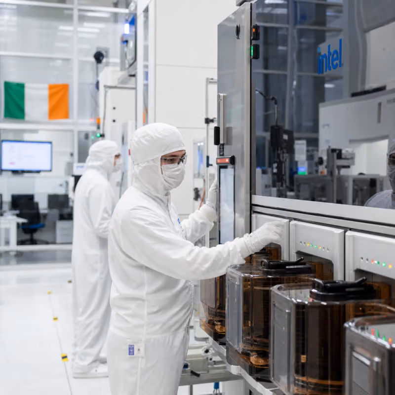 Technicians in cleanroom suits at a wafer fabrication tool bearing the Intel logo.